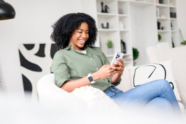 Neşeli bir Afro-Amerikalı kadın rahat bir şekilde koltuğunda oturuyor, akıllı telefonuna dalmış, eğlenceli bir videonun ya da keyifli bir sohbetin tadını çıkarıyor, parlak, çağdaş bir yaşam alanında.