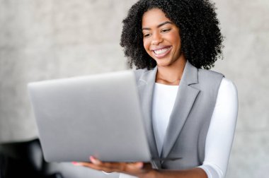 Profesyonel bir Afro-Amerikalı iş kadını dizüstü bilgisayarını tutarken özgüvenle ışıldıyor, muhtemelen başarılı bir anlaşmayı tartışıyor. İş ortamında başarı ve güvenin tasviri