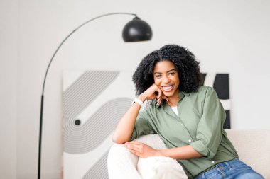 Kanepede dinlenen Afro-Amerikan bir kadın büyüleyici bir gülümseme ve elleri yanağında, minimalist bir oturma odasında, huzurlu ve modaya uygun bir ev sahnesini gösteren rahatlatıcı bir titreşim yayıyor.