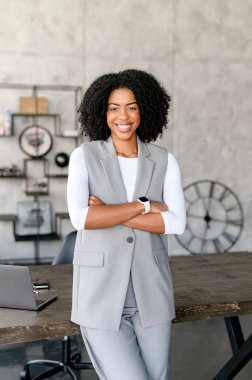 Kendinden emin bir Afro-Amerikalı iş kadını modern ofis ortamında kollarını kavuşturmuş, gülümsemesi profesyonellik ve yakınlık saçıyor. Başarılı, güçlü bir girişimci.