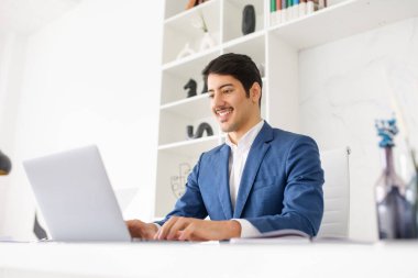 Profesyonel bir İspanyol işadamı masasında oturmuş laptopa odaklanmış vaziyette iş dünyasının karmaşıklıklarının yönetimindeki rolünü vurguluyor.