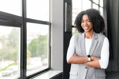 Afro-Amerikalı bir iş kadını modern bir ofiste kollarını kavuşturarak duruyor. Büyük pencereler ardında, kendine güvenen gülüşü ve dengeli duruşu modern kadın liderliğinin güçlü bir imajını yansıtıyor..