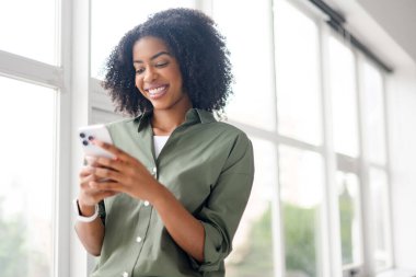 Afro-Amerikalı bir kadın akıllı telefonuna mesaj atıyor, gülüşü bir arkadaşıyla veya sevdiği biriyle hoş bir alışverişi ima ediyor, dijital bağlantının sevincini gösteriyor..