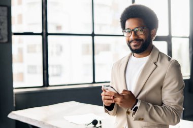 Kendine güveni ve cazibesi ile, akıllı bej takım elbiseli genç Afro-Amerikan işadamı telefonunu kontrol ediyor, modern iş hayatının dinamik temposunu somutlaştırıyor, akıllı telefon kullanan erkek girişimci