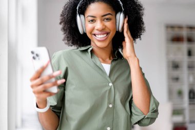 Neşeli bir Afro-Amerikalı kadın akıllı telefonuyla büyük kulaklıklar takıyor, minimalist ve zarif bir ev ofisine karşı kuruluyor, moda ve bağlantılı bir yaşam tarzı yayıyor..