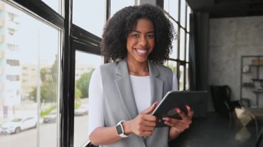 Profesyonel kıyafetli ışıl ışıl bir Afro-Amerikalı kadın tabletine gülümsüyor, şehir manzarası arkasında büyük bir pencereden, modern iş ve kentsel yaşam tarzının bir karışımını canlandırıyor..