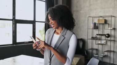 Afro-Amerikalı iş kadını, samimi bir neşe anında, pencerenin kenarındaki akıllı telefonuna göz gezdirirken, kendinden emin, teknoloji meraklısı varlığının altını çizen doğal ışıkla yıkanırken yakalandı..