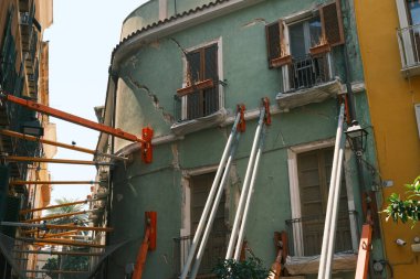 Görünür çatlakları olan, iskele destekli sıkıntılı bir binayı gösteren bir resim. Canlı yeşil cephe çevre mimariyle çelişiyor ve kentsel restorasyon çabalarını vurguluyor.