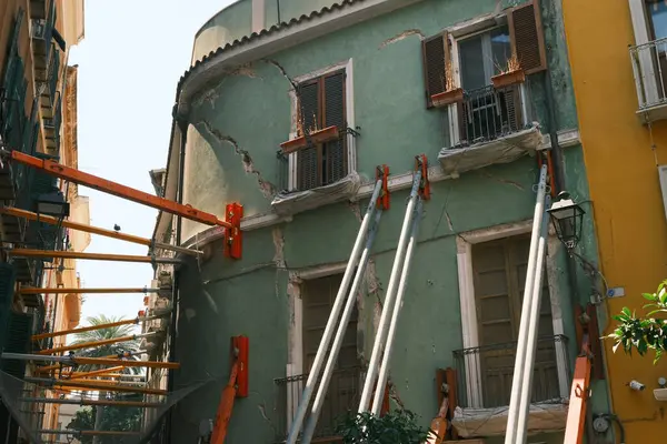 Görünür çatlakları olan, iskele destekli sıkıntılı bir binayı gösteren bir resim. Canlı yeşil cephe çevre mimariyle çelişiyor ve kentsel restorasyon çabalarını vurguluyor.