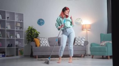 cheerful young woman in headphones dances with mop in hands while cleaning floors at home