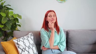 thoughtful woman with red hair thinks about solving a problem while sitting on sofa in room