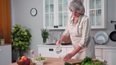 portrait of a elderly woman pouring a glass of cool healthy water while standing in kitchen, smiling and looking at camera