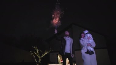 family party, joyful woman and man with their children have fun launching fireworks against the backdrop of a house with bright lights in the late evening
