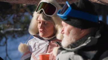 recreation of old husband and wife, romantic elderly couple drinking hot tea in thermos during winter walk in the forest backdrop of snow
