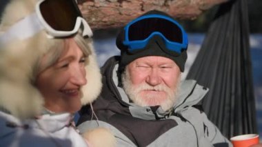 romantic vacation of elderly spouses, an old man communicates with wife drinks hot tea from a thermos sitting on a hammock in a winter forest