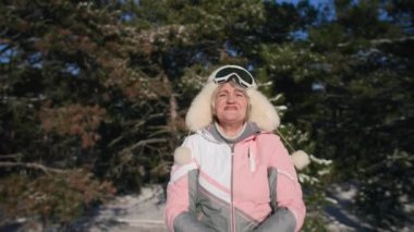 winter outdoor recreation, elderly woman in warm winter clothes shines snow in sunny weather while relaxing in forest, smiling and looking at camera
