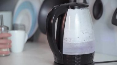 electric kettle with hot water on background of women making coffee and pouring sugar into cup