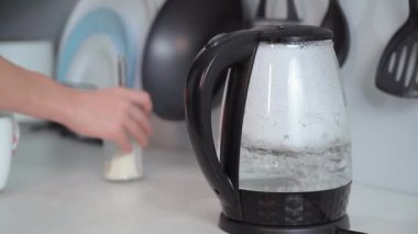 young woman pouring hot water from an electric kettle into cup on background of kitchen
