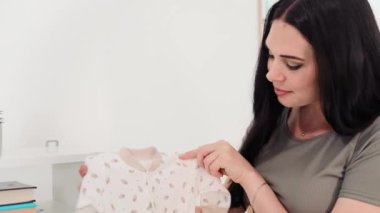 Hamile kadının portresi yeni doğan için nazikçe kıyafet katlıyor, gülümsüyor ve kameraya bakıyor.