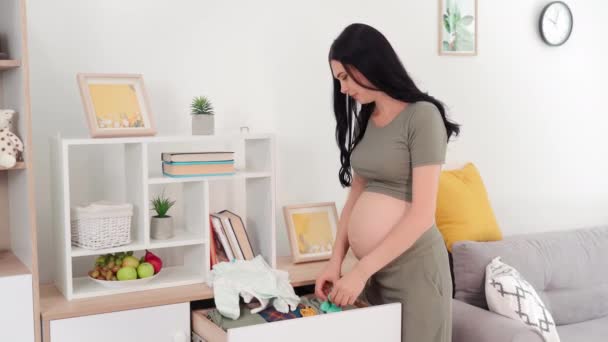 在母亲的指导下 年轻的孕妇在家里把天然面料新生儿的衣服放进抽屉的箱子里 为婴儿的出生做准备 — 图库视频影像
