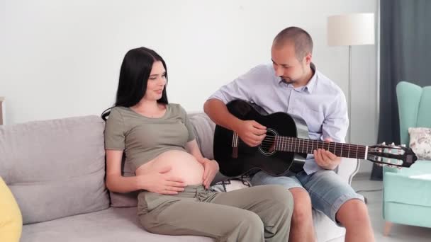 为人父母 有爱心的年轻父亲为胎儿在子宫里弹奏吉他 妻子坐在家里的沙发上 — 图库视频影像