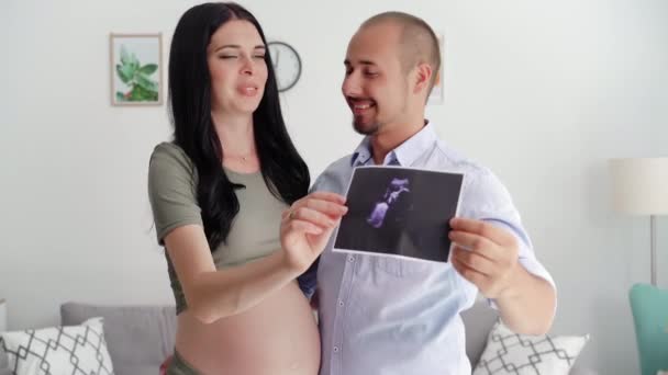 年轻男子和怀孕妻子的画像 手里拿着超声照片 靠着白墙站着 — 图库视频影像