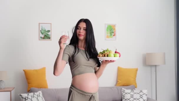 怀孕期间适当的营养 在舒适的房间里摆出怀孕妇女的画像 拿着一盘水果和一杯清洁的水 微笑着看着镜头 — 图库视频影像