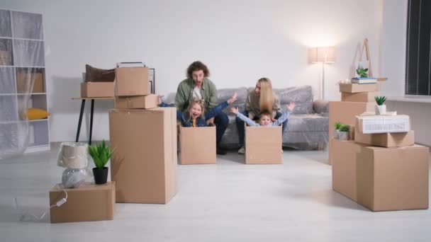 新しい家に引越しの日の休日男性と女性の両親は家具の間で新しいアパートの床にそれらを転がす箱の中で小さなかわいい子供と楽しいです — ストック動画