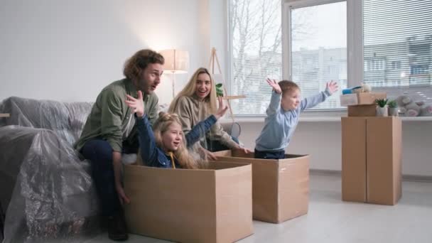 新しい家に移り住む若い母親と父親は家具を背景に箱の中で娘と息子を転がすのが楽しい — ストック動画