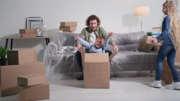 若いお父さんと娘はソファに座ってる間に荷解きしてる可愛い男の子供が新しいアパートに引っ越してる間に箱から飛び降りて不動産 — ストック動画