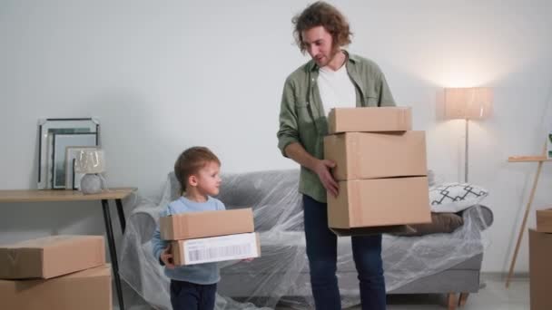 不動産屋で小さな魅力的な息子を持つ笑みを浮かべて箱を手にした若い男は家具の背景に立って新しいアパートに移動することを喜んで — ストック動画