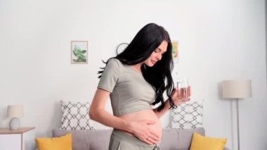 Çekici hamile kadın göbeğine elleriyle dokunur ve bardakta temiz su içer, gülümser ve kameraya bakar.