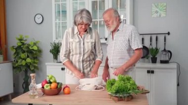 Sağlıklı ürünler, çekici yaşlı bir kadın ve yaşlı bir adam çevre dostu bir alışveriş çantası hazırlıyorlar ve mutfakta salata yapmak için taze sebzeler çıkarıyorlar.
