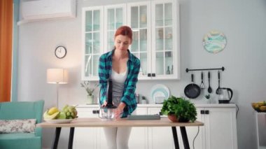 Genç bir kadın meyve püresi yapmak için elektrikli blender kullanıyor. Mutfakta masada dururken fişe takıyor.