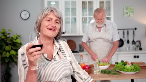 台所での夕食のためのサラダを切断夫のワインの背景のガラスとエプロンで喜びの老婦人の肖像画 笑顔とカメラを見て — ストック動画