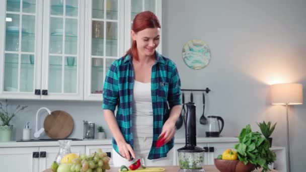 快乐的女人在厨房的餐桌边 微笑着看着镜头 观察着适当的营养 并在新鲜的蔬菜面包中准备了一个冰沙 — 图库视频影像