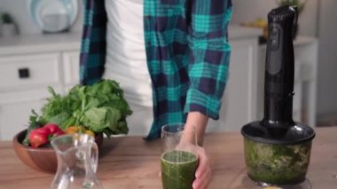portrait of a smiling young woman holding a glass of fresh vegetable smoothie in hands, taking a sip smiling and looking at camera