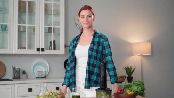 一个年轻女子的画像 她站在厨房桌子旁边 手里拿着搅拌机和新鲜蔬菜 一边做着一个光滑的面庞 一边微笑着 一边看着相机 — 图库视频影像