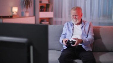 Kablosuz joystick 'i olan modern yaşlı adam akşam evde dinlenirken kanepede otururken TV' de video oyunları oynuyor.