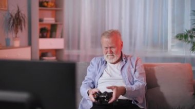 Elinde kablosuz joystick olan çekici bir emekli, kanepede otururken TV 'de video oyunları oynuyor, akşamları rahat bir odada dinleniyor.