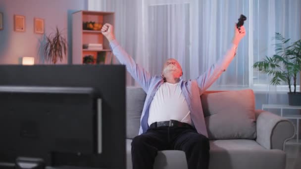 轻松愉快的可爱老人坐在客厅的沙发上 用无线游戏机在电视机上玩电子游戏 — 图库视频影像