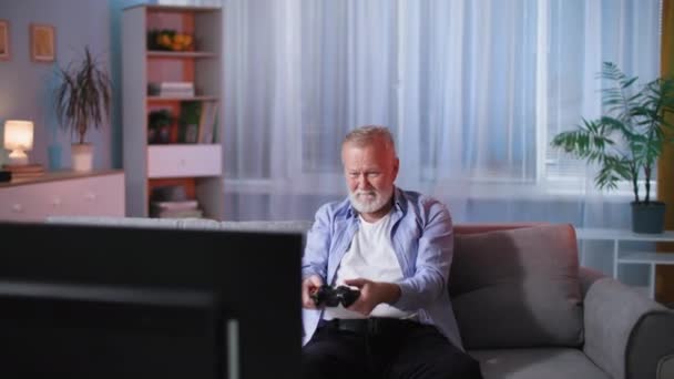 在家里的房间里 一个老人坐在电视机前的沙发上 一边玩着电子游戏 一边玩着游戏 — 图库视频影像