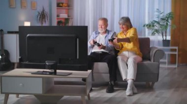 Yaşlı erkek ve kadın oyuncular akşamları oturma odasındaki kanepede otururken, oyun konsolu ve joypad 'i olan bir TV' de modern teknoloji kullanır ve video oyunları oynarlar.