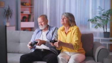 Mutlu, evli bir çift oyun konsolu ve televizyonla oynayan kameramanlarla eğlenirken kanepede oturup gülümsüyor ve kameraya bakıyor.