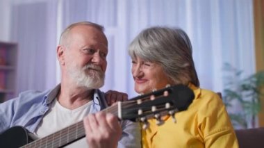 Yaşlı ve romantik bir çift birlikte gitar çalarak ve kanepede oturup kameraya bakarak şarkı söyleyerek eğleniyor.