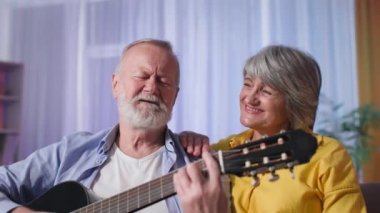 Gülümseyen eş, evde dinlenirken akustik gitar çalan kocasına şarkı söyler ve kameraya bakar.