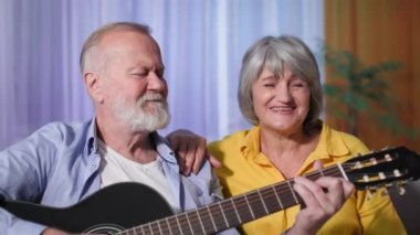 Yaşlı erkek ve kadın emeklilerin evdeki kanepede oturmuş, gülümseyip kameraya bakarken gitar çalarak ve şarkı söyleyerek eğlenen portreleri.