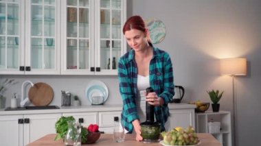 Çekici bir kadının portresi taze sebzelerden ve blender 'da ezilmiş marullardan sağlıklı vejetaryen smoothie hazırlarken mutfakta durup bardaktan kokteyl yudumlarken, gülümserken ve bakarken.