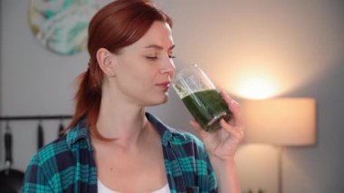 Elinde taze sebze ve maruldan bir bardak yeşil smoothie tutan, bir yudum alıp gülümseyen ve kameraya bakan tatlı bir kadının portresi.