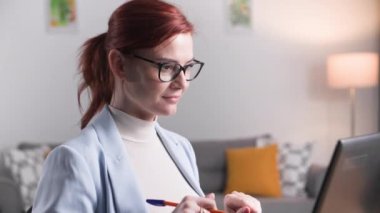 Genç bir kadın, odada dizüstü bilgisayarının başında otururken, düşünceli bir şekilde gözlüklerini takıp işini yapıyor.
